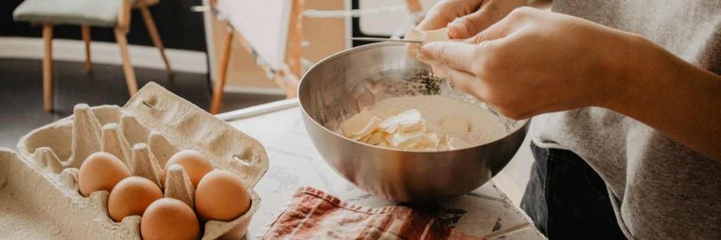 A person baking at a kitchen bench. They are mixing in eggs, flour, and white chocolate in a bowl. 