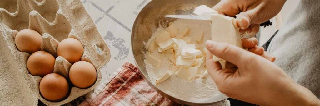 A person baking. Eggs are on the kitchen bench, flour and butter in a bowl, flour on the bench and a person mixing the ingredients. 