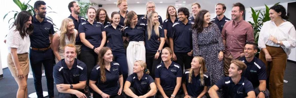 TRS new graduate team 2021 in a large room posing for a group photo. Mix of men and women all wearing navy blue TRS polo's. Some are seated on the floor and the others are standing behind them. 