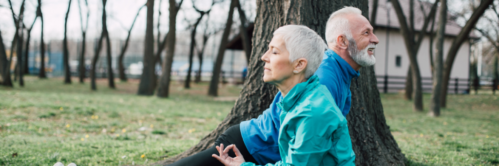 Older couple, a man and a woman both stretching and meditating. They have blue jumpers and black long pants on and have grey hair. 