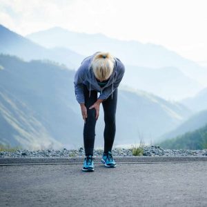 A lady with blonde hair bending over grabbing at her knee. She has a blue jacket and long black leggings and is running in a mountain trail