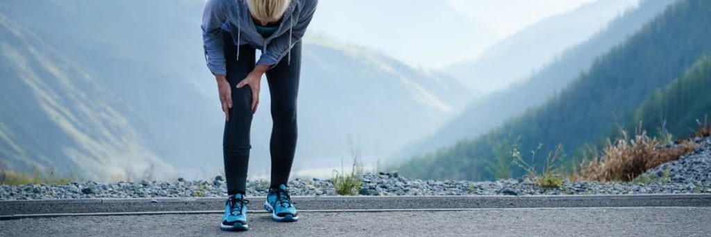 A lady with blonde hair bending over grabbing at her knee. Wearing long black tights and a blue spray jacket. Mountains and paved road in the background 